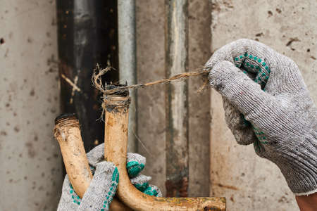 Two pieces of an old rusty bent plumbing pipe that has become unusable due to corrosion, a plumber in work gloves shows the time in the process of being replaced with modern communicationsの写真素材