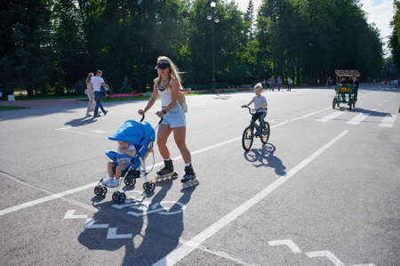 Saint Petersburg, Russia - August 18, 2020: A girl on roller skates rides with a child in a baby carriage in Victory Park on a summer sunny day on a bicycle path on the central alleyのeditorial素材