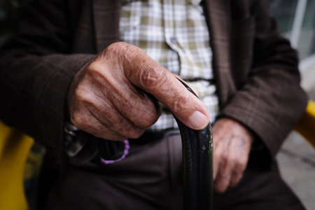 An elderly unrecognizable man sits on a chair with a stick in his hands. Wrinkled hands close up. Conceptual photography symbol about aging and old ageの写真素材