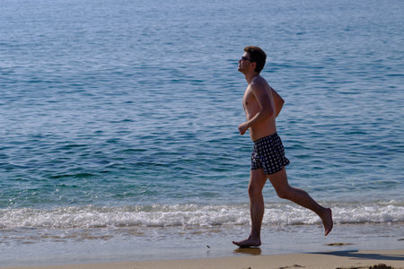 Young handsome man 25-30 years old, brunette, in weed sunglasses, makes an easy run along the sandy sea coast, naked torso sunglassesの写真素材