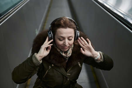 A pleasant blonde 35-40 years old inside the subway listens to music through high-quality over-ear headphones connected to the phone via bluetooth.の写真素材