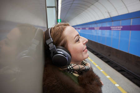 A pleasant blonde 35-40 years old inside the subway listens to music through high-quality over-ear headphones connected to the phone via bluetooth.の写真素材