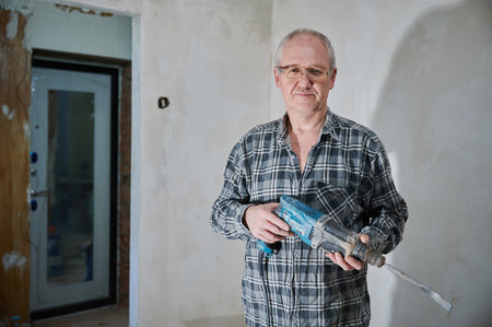 Portrait of a man 55-60 years old in a working environment during the repair of an apartment with his own hands, in a plaid shirt, smiling, wearing glasses, holding a perforatorの写真素材
