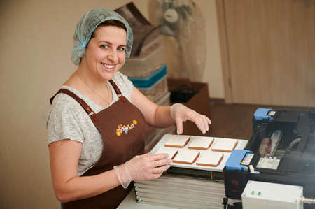 Pleasant middle-aged woman in the process of printing images on gingerbread with food coloring. Small wholesale production line, manufacturing processの写真素材