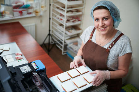 Pleasant middle-aged woman in the process of printing images on gingerbread with food coloring. Small wholesale production line, manufacturing processの写真素材