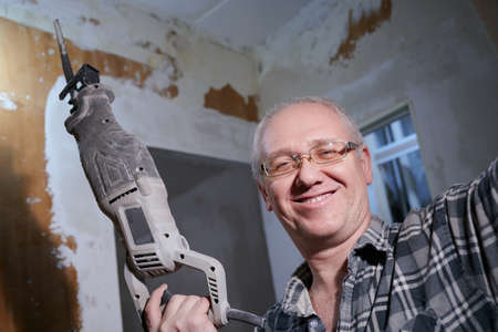 Portrait of a man 55-60 years old in a working environment during the repair of an apartment with his own hands, in a plaid shirt, smiling, wearing glasses, holding a perforatorの写真素材
