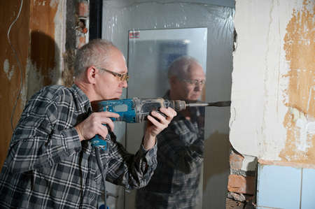 Portrait of a man 55-60 years old in a working environment during the repair of an apartment with his own hands, in a plaid shirt, smiling, wearing glasses, holding a perforatorの写真素材