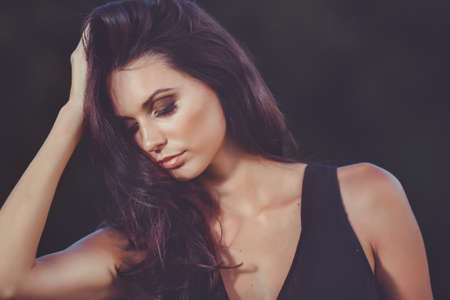 Portrait of a serious brunette girl in black dress closeup with a hand on hair on black backgroundの写真素材