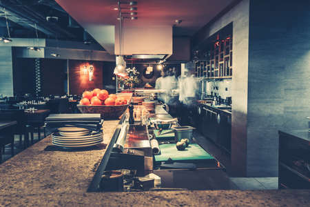 Restaurant kitchen interior: bar counter made of natural stone, fences off the open kitchen and hall for visitors with tables and chairs. In the background buzzing restaurant work motion chefsの写真素材