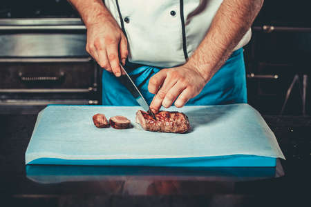 Chef in hotel or restaurant kitchen dressed in blue apron cooking, only hands, he is cutting meat or steak with big sharp knifeの写真素材