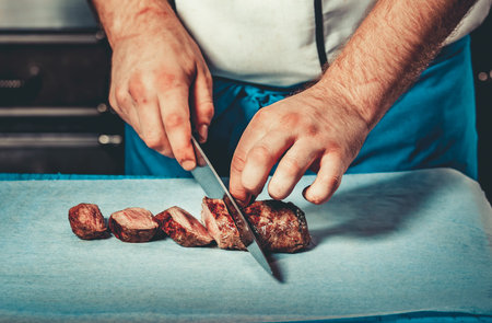 Chef in hotel or restaurant kitchen dressed in blue apron cooking, only hands, he is cutting meat or steak with big sharp knifeの写真素材