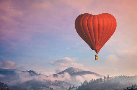 Beautiful red air balloon heart shape against blue and pink pastel sky in a sunny bright morning. Foggy mountains in the background. Romantic trip on Valentines Day. Sport and recreation travel themeの写真素材