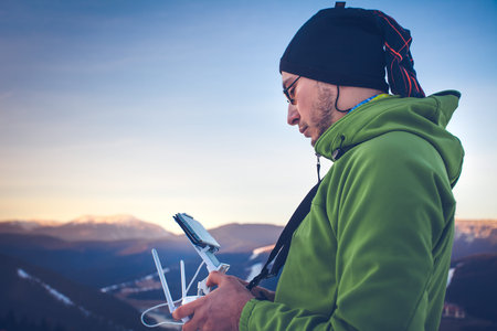 Young man in green jacket operating a drone using a remote controller. Ski resort in the background,...