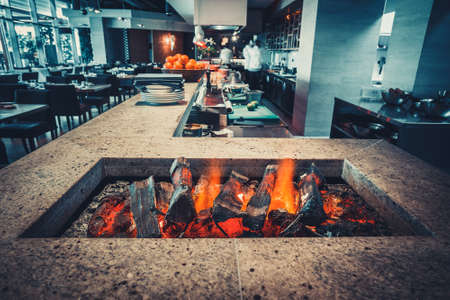 Restaurant kitchen interior: brazier with burning wood, made of natural stone with fire for BBQ. In the background buzzing restaurant work motion chefsの写真素材