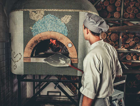 Food concept. Preparing traditional italian pizza. Young chef in white uniform holds in his hands shovel for pizza, he bakes dough in a professional oven in interior of modern restaurant kitchenの写真素材