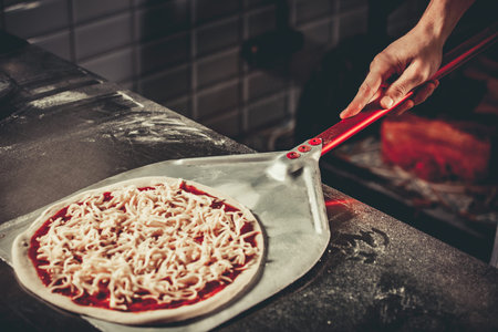 Food concept. Preparing traditional italian pizza. Chef holds long irob shovel for pizza, baking dough with pasta and cheese in a professional oven. Only hands close upの写真素材