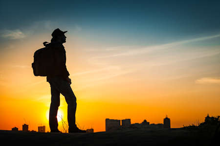 Traveler silhouette watching amazing sunset. Young casual man with backpack and cowboy hat standing alone on hill above evening cityscape. Lifestyle Travel Concept Outdoor Background. Kiev, Ukraineの写真素材