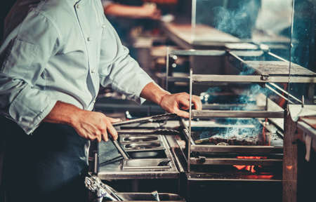 Food concept. Chef in white uniform monitors the degree of roasting and turns meat with the forceps in interior of restaurant kitchen. Preparing traditional beef steak on barbecue oven. Only handsの写真素材