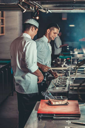 Food concept. Young handsome chefs in white uniform kindle coals and monitor temperature degree for roasting meat in interior of restaurant kitchen. Preparing traditional beef steak on barbecue oven.の写真素材