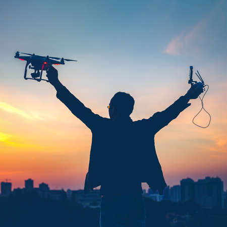 Silhouette of man holding switched on Drone quad copter and Remote control enjoying freedom, victory, success. Cityscape with dramatic sunset sky in the background. Business concept, raised handsの写真素材