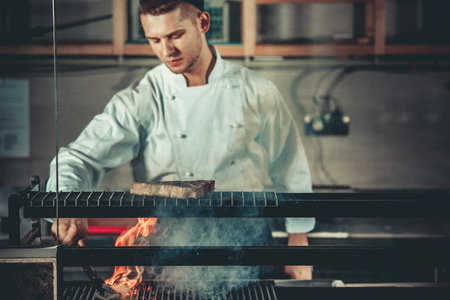 Food concept. Young handsome chef in white uniform monitors the degree of roasting and turns meat with the forceps in interior of restaurant kitchen. Preparing traditional beef steak on barbecue oven.の写真素材