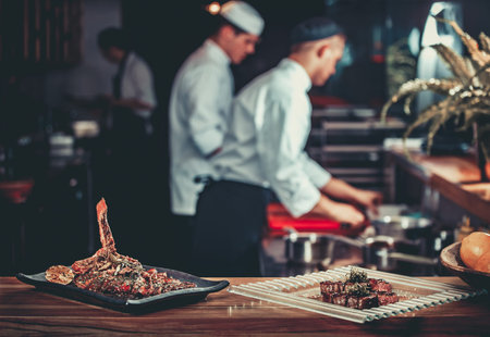 Food concept. Ready grilled pork ribs dish and beef steak with herbs. Ready to serve. Ready to eat. Two chefs working in the background interior of modern professional restaurant kitchenの写真素材