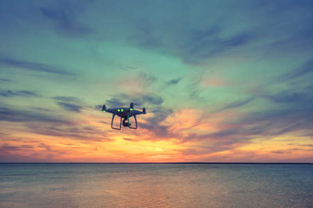 Silhouette of Drone Quad copter flying in sky. Aerial panoramic view of sunset over ocean. Dramatic picturesque evening scene. Colorful cloudy sky in the background. Vehicle at sundown and copy spaceの写真素材