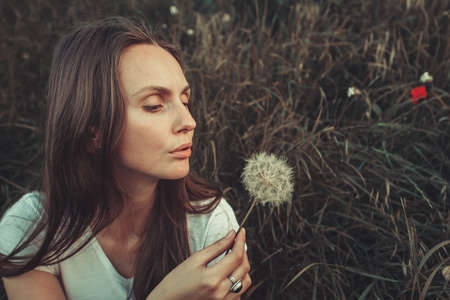 Beautiful Young Woman sitting in green grass and blowing dandelion. Outdoors. Enjoy Nature. Healthy Caucasian Girl on spring lawn. Allergy free concept. Vintage filterの写真素材