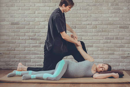 Traditional Thai Massage of pregnant woman lying on the mat in yoga studio. Young white masseur dressed in a black uniform stretches her hand. Brick wall in the background. Health, relax, spa conceptの写真素材