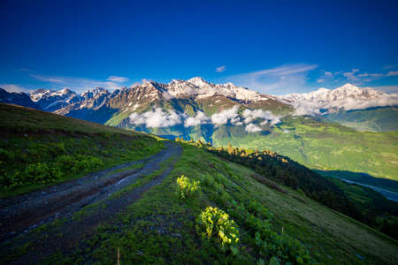 Panoramic summer landscape with green hill, mountain snow capped peaks and trekking path against blue clear sky. Svanetia region, Georgia. Main Caucasian ridge. Travel background.の写真素材