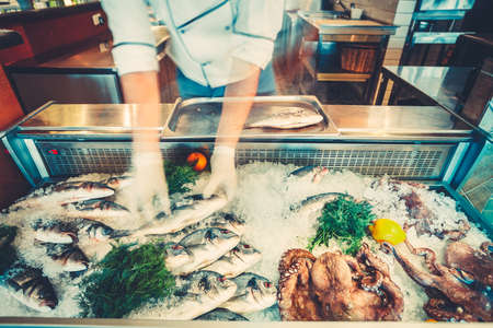 The chef-cooker is taking the freshly caught fish out of the refrigerated counter for the cooking delicious meal in the restaurant. Fresh seafood the fish and octopuses for healthy lifestyle.の写真素材