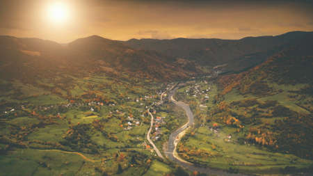 Aerial View: Beautiful canyon village located along the river. Dramatic autumn mountain landscape with green meadows, orange pine tree forests, sunset cloudy sky. Carpathians, Ukraine, Europe.の写真素材