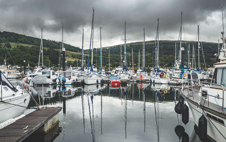 Numerous yachts at the Northern Ireland pier. Rainy cloudy sky background. Small touristic ships at the bay. Reflection in the water surface. Luxury lifestyle. Green hills before the rain.の写真素材