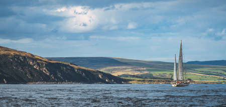 Panoramic view of Irish shoreline. Lone sailing yacht among the calm sea water. Northern Ireland. Blue cloudy sky background. The sunset over the Irish land. Picturesque scenery. Outdoor activity.の写真素材