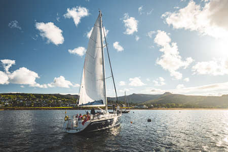 Tourists on the sailing yacht. Northern Ireland. Breathtaking marine scenery. The lone boat on the water surface. Irish shoreline on the horizon. Sunset blue cloudy sky. Beauty of Irish nature.の写真素材