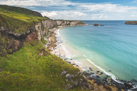 White sand beach of the Northern Ireland shoreline. Green grass covered cliff washed by the turquoise transparent sea water. Beauty of wild untouched environment. Breathtaking Irish landscape.の写真素材
