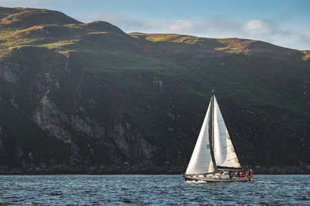 Touristic yacht sailing next to the Northern Ireland steep rocks. Green covered heels, shoreline. Outdoor activity. Perfect background for the creating of various kinds of collages and illustrations.の写真素材