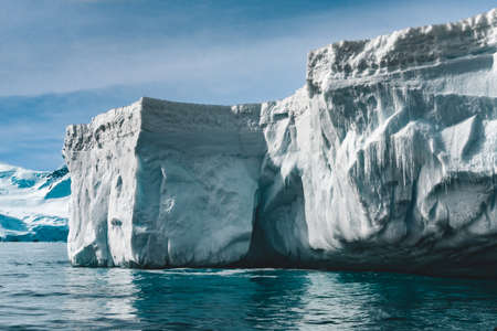 Close-up sunlit iceberg. Antarctica scene in blue and white tints. Amazing snow covered block of ice with icicles floating among the polar ocean. The cloudy sky background. Picturesque winter scenery.の写真素材