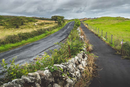 Close-up countryside roads divided by the stone boulevard. Islay island, Scotland. The route to the Laphroaig distillery, Green grass covered fields surrounding. The dark cloudy sky before the rain.の写真素材
