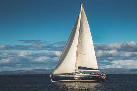 Close-up sailing yacht. Northern Ireland. Outdoor activity. Lone boat on the water surface. Overwhelming marine scene. Irish shoreline on the horizon. Blue cloudy sky background.の写真素材
