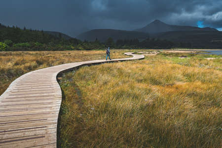 Tourist walking on the wooden path. Northern Ireland. Mighty mountains background. Rainy cloudy sky in grey tints. The backpacker on the trail. Hiking among the wild environment. Outdoor activity.の写真素材