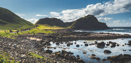 Tourist path next to the Northern Ireland shoreline. Amazing Irish landscape. Green covered hills under the blue cloudy sky. Boulders beach. Outdoor hiking activity. Tourist attraction.の写真素材