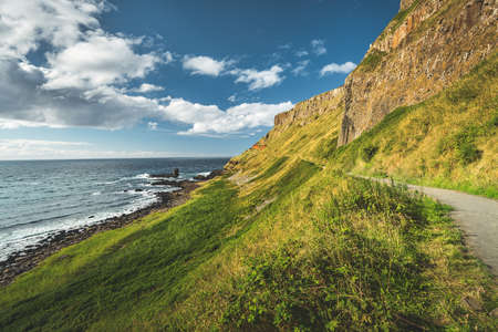 Steep green slope with tourist path. Northern Ireland. The grass covered hill next to the ocean. Overwhelming Irish shoreline scene. Blue cloudy sky background. Perfect touristic site.の写真素材