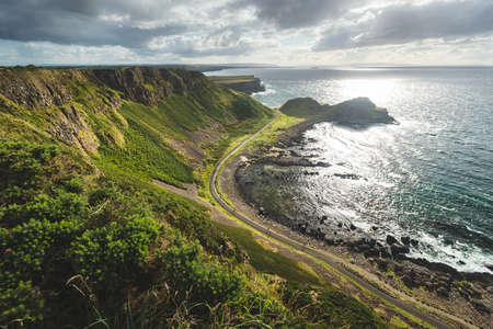 Sunlit ocean, green shore with the road. Northern Ireland landscape. Stunning lowland next to the ocean water. Panoramic overview of the untouched nature. Cloudy sky above the Irish shoreline.の写真素材