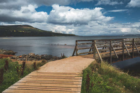 Close-up wooden pier connecting the sea and grass covered land. Northern Ireland. Stunning shoreline among the wild virgin nature. Blue cloudy sky background. Amazing Irish landscape.の写真素材
