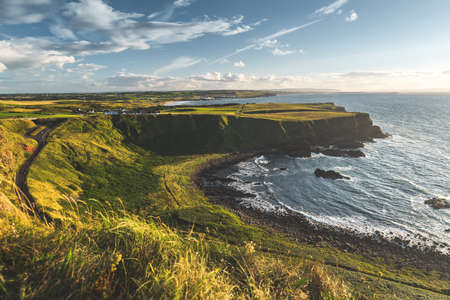 Sunlit Northern Ireland shoreline. The green grass covered land next to the ocean water surface. The local road among the wild untouched nature. The stunning bay under the blue cloudy sky. Serenity.の写真素材