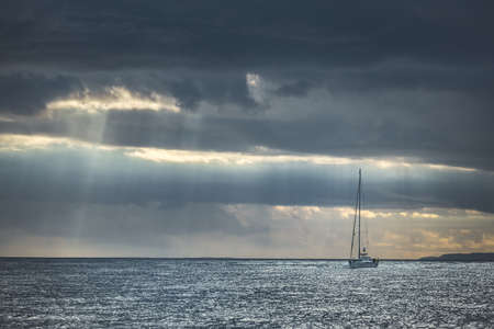 Rainy cloudy sky above the lone yacht sailing in the sea. Northern Ireland. Sun lights, rain flows over the sea water. Lonely boat among the ocean. Overwhelming marine landscape in blue grey tints.の写真素材