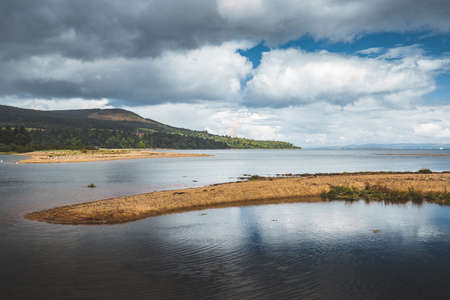 Piece of land among the calm sea. Northern Ireland. The sand pier surrounded by the serene water surface. Overwhelming Irish landscape. Rainy sky background. Beauty of wild Irish nature. Peacefulness.の写真素材