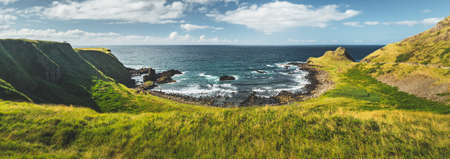 Panoramic overview of the Northern Ireland shoreline. Amazing Irish landscape. Green grass covered shore surrounded by the ocean water. Cozy bay under the cloudy sky background. Wilderness.の写真素材
