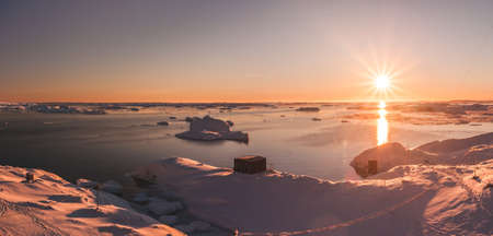 Bright orange sunset over the Antarctica shoreline and Vernadsky station. Amazing panoramic view of the sunlit polar bay. The snow covered surface of the South Pole next to the frozen water surface.の写真素材